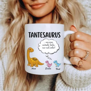 May include: White ceramic mug with the text "TANTESAURUS" and a cartoon dinosaur illustration. The mug features a thought bubble with German text and the names "Anna", "Emilia", and "Noah" below the dinosaurs.