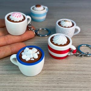 Puede incluir: Cinco tazas de cerámica en miniatura con rayas de diferentes colores y coberturas, incluyendo crema batida, chispas y malvaviscos, unidas a llaveros. Las tazas están dispuestas sobre una superficie de madera.