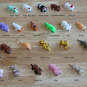 May include: A collection of 24 colorful animal figurines, including a Christmas hippo, a polar bear, a panda, a reindeer, a cat, an elephant, a duck, a clownfish, a chameleon, an axolotl, a hammerhead shark, a grizzly bear, a goldfish, a giraffe laying down, a giraffe sitting, a gecko, a fox, a triceratops, a sloth, a puppy dog, a pig, an otter, and a hippo.