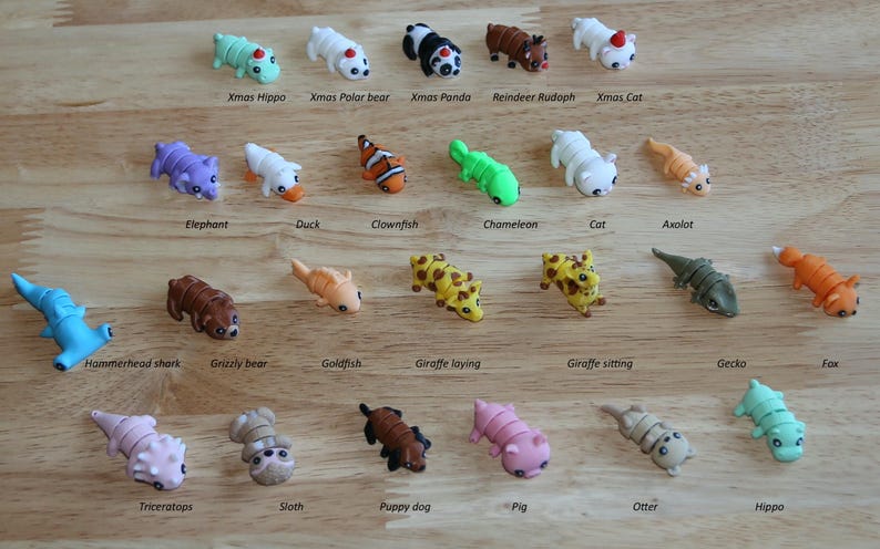 May include: A collection of 24 colorful animal figurines, including a Christmas hippo, a polar bear, a panda, a reindeer, a cat, an elephant, a duck, a clownfish, a chameleon, an axolotl, a hammerhead shark, a grizzly bear, a goldfish, a giraffe laying down, a giraffe sitting, a gecko, a fox, a triceratops, a sloth, a puppy dog, a pig, an otter, and a hippo.