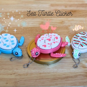 May include: Three colorful sea turtle clickers with different patterns. The blue turtle has a snowflake pattern, the pink turtle has a heart pattern, and the white turtle has a green and red striped pattern. Each turtle has a small chain attached.