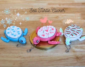 May include: Three colorful sea turtle clickers with different patterns. The blue turtle has a snowflake pattern, the pink turtle has a heart pattern, and the white turtle has a green and red striped pattern. Each turtle has a small chain attached.