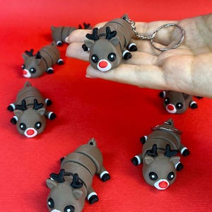 May include: A set of six brown and black reindeer keychains with red noses. The reindeer are segmented and can be bent into different positions. The keychains are on a red surface.