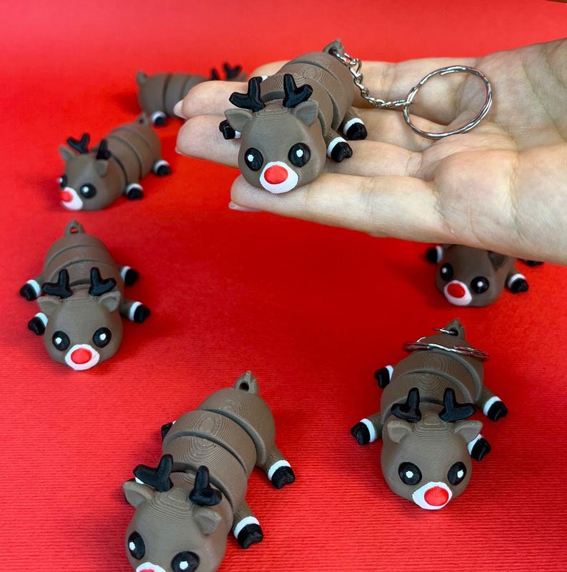 May include: A set of six brown and black reindeer keychains with red noses. The reindeer are segmented and can be bent into different positions. The keychains are on a red surface.