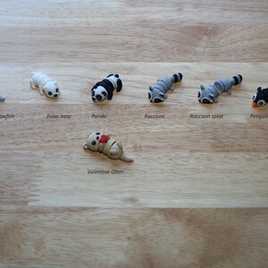 May include: A set of seven small animal figurines, including a sawfish, polar bear, panda, raccoon, raccoon split, penguin, and a Valentine's Otter. The figurines are all made of a light gray material and are arranged in a row on a wooden surface.