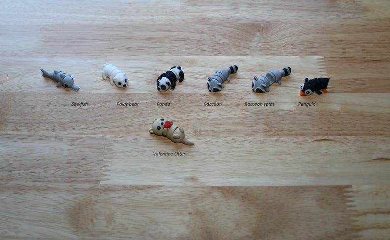 May include: A set of seven small animal figurines, including a sawfish, polar bear, panda, raccoon, raccoon split, penguin, and a Valentine's Otter. The figurines are all made of a light gray material and are arranged in a row on a wooden surface.
