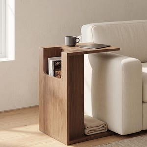 May include: A C-shaped wooden side table with a dark brown finish, featuring a vertical storage compartment for books and a lower shelf for a folded blanket. The tabletop holds a grey mug and a tablet, positioned next to a white upholstered sofa.