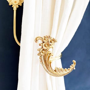 Könnte beinhalten: Vergoldete Gardinenhalter mit floralen und blattartigen Verzierungen. Einer hält einen weißen Vorhang, der andere ist an einer dunkelblauen Wand befestigt. Die Halterungen im Vintage-Stil verleihen eine dekorative Eleganz.