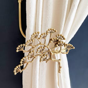 May include: A gold-coloured metal curtain tieback shaped like a tree branch with small, round buds. The tieback holds a white curtain against a dark blue wall. Another gold-coloured curtain rod holder is visible.