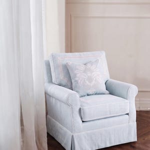 May include: A light blue upholstered armchair featuring white piping along the edges, a skirted base, and a square throw pillow with a white floral damask pattern centered on the back cushion. The chair has rolled arms and a structured back.
