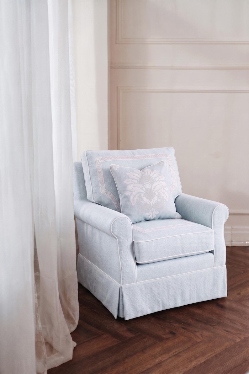 May include: A light blue upholstered armchair featuring white piping along the edges, a skirted base, and a square throw pillow with a white floral damask pattern centered on the back cushion. The chair has rolled arms and a structured back.