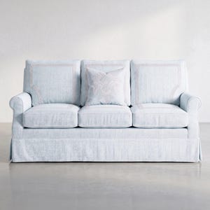 May include: A light blue three-cushion sofa featuring a skirted base, rolled arms, and three back cushions with a subtle rectangular border pattern. A single square throw pillow with a central floral motif rests against the middle back cushion.