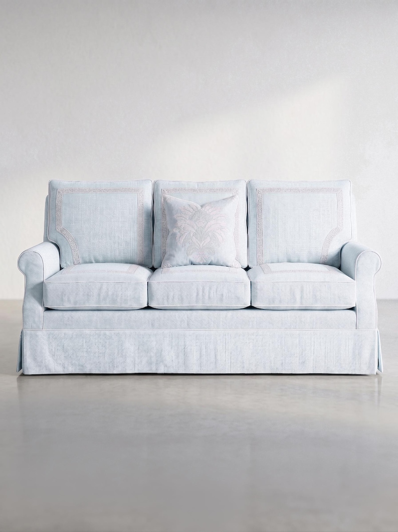 May include: A light blue three-cushion sofa featuring a skirted base, rolled arms, and three back cushions with a subtle rectangular border pattern. A single square throw pillow with a central floral motif rests against the middle back cushion.