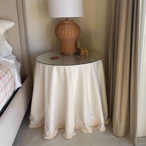 May include: A white table skirt with a fringe trim sits on a round glass table top. A wicker lamp with a white lampshade sits on the table. The table is next to a bed with a white headboard and a pink and white plaid blanket.