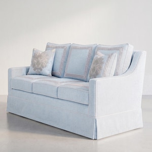 May include: A light blue sofa featuring a skirted base, white piping trim, and three back cushions with rectangular border detailing. Two square throw pillows with a centered beige floral pattern are placed against the back cushions.