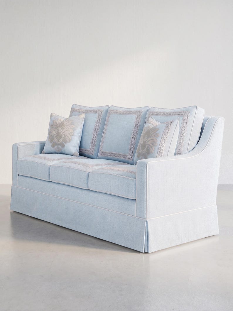 May include: A light blue sofa featuring a skirted base, white piping trim, and three back cushions with rectangular border detailing. Two square throw pillows with a centered beige floral pattern are placed against the back cushions.