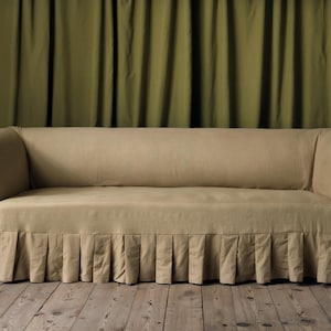 以下が含まれることがあります： 緑色のカーテンの前に置かれた、プリーツスカートと丸みを帯びた肘掛けが付いたベージュのソファです。