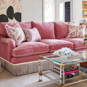 May include: A pink velvet sofa with white fringe trim and three pillows. The sofa is in a living room with a glass coffee table and books on the lower shelf.