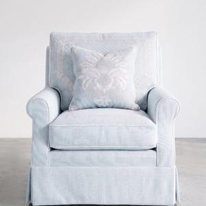 May include: A light blue armchair with white piping and a skirted base. The chair features a square backrest with a Greek key border pattern and a matching square throw pillow centered on the seat, decorated with a large, pale floral damask motif.