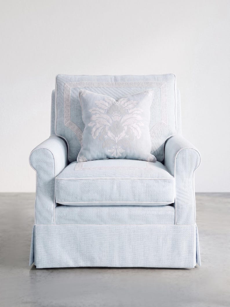 May include: A light blue armchair with white piping and a skirted base. The chair features a square backrest with a Greek key border pattern and a matching square throw pillow centered on the seat, decorated with a large, pale floral damask motif.