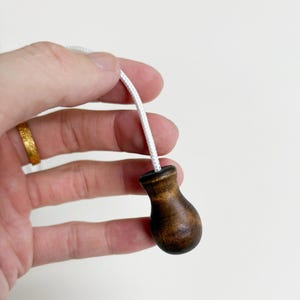 May include: A wooden light pull with a dark brown, teardrop-shaped handle. The handle is attached to a white cord. The pull is held against a white background.