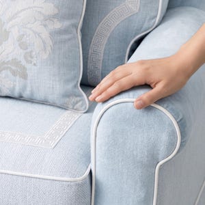 May include: A light blue upholstered armchair featuring white piping along the seams and a white Greek key pattern border. A matching light blue throw pillow with a white floral damask design is positioned against the backrest of the chair.