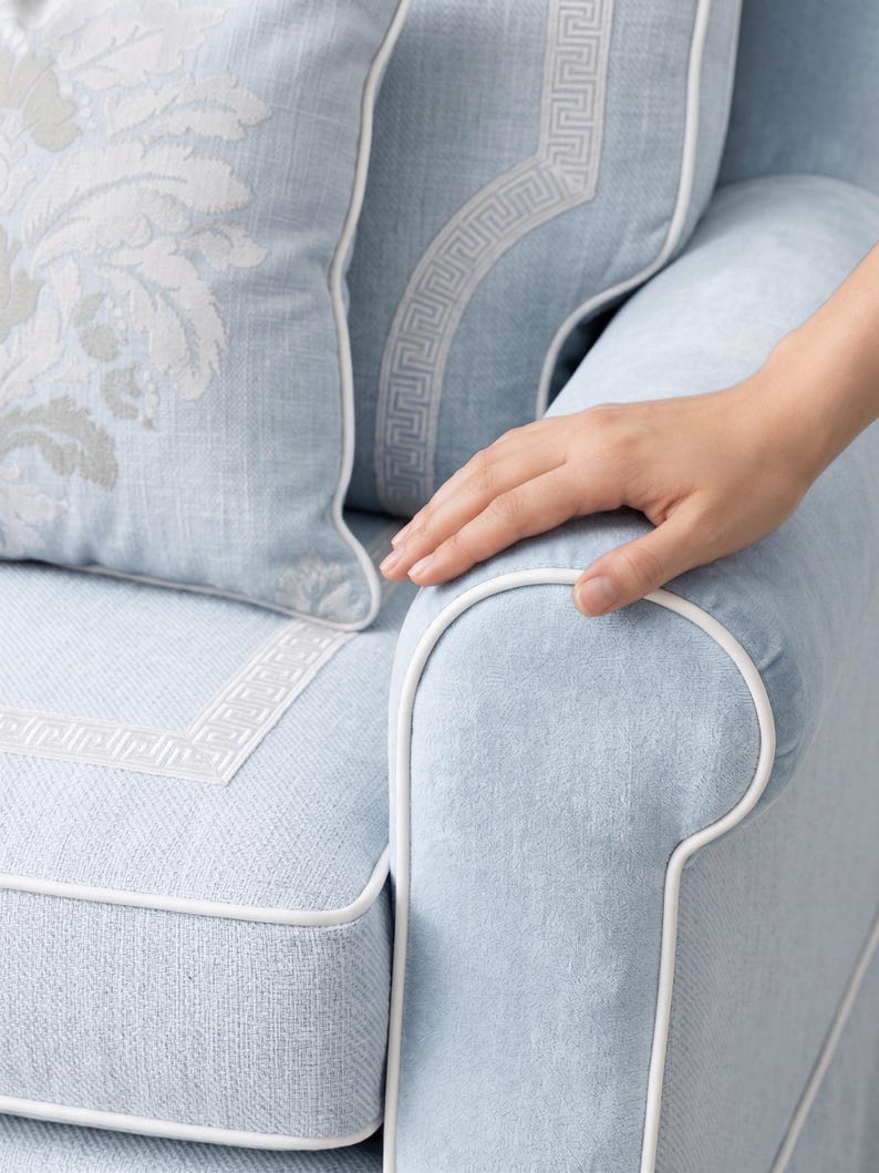 May include: A light blue upholstered armchair featuring white piping along the seams and a white Greek key pattern border. A matching light blue throw pillow with a white floral damask design is positioned against the backrest of the chair.
