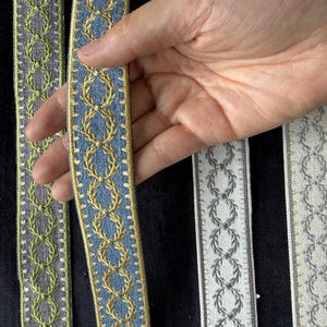 May include: Five strips of decorative woven trim featuring a repeating laurel wreath pattern. The colors include orange, lime green, blue with gold, grey, and cream with silver. Each strip has a distinct border and textured fabric finish.