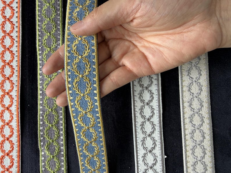 May include: Five strips of decorative woven trim featuring a repeating laurel wreath pattern. The colors include orange, lime green, blue with gold, grey, and cream with silver. Each strip has a distinct border and textured fabric finish.