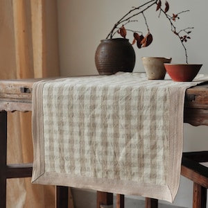 Puede incluir: Un camino de mesa a cuadros beige y blanco con un borde marrón descansa sobre una mesa de madera rústica. Un jarrón marrón, dos cuencos de cerámica pequeños y una rama con hojas secas están dispuestos sobre la mesa.