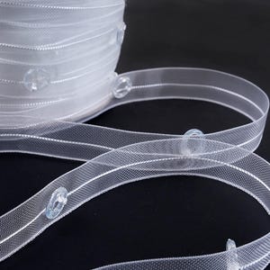 May include: A roll of white, sheer elastic ribbon with clear, oval-shaped buttons. The ribbon has two thin, white stripes running lengthwise. The buttons are evenly spaced along the ribbon, which is likely used for sewing or crafting projects.