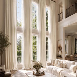 May include: Floor-to-ceiling off-white curtains hang from a dark metal rod, covering tall arched windows. The fabric features vertical pleats that extend from the top to the floor, creating a uniform, structured appearance across the window wall.
