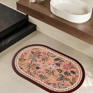May include: An oval-shaped bathroom rug featuring a soft peach-colored background with a detailed botanical print of various flowers and tropical leaves. The rug is framed by a decorative cream-colored border and a solid dark burgundy outer edge.