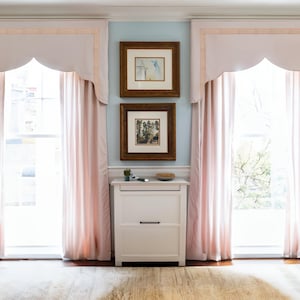Op de afbeelding: Twee ramen met roze gordijnen en lambrequins, ingelijste kunstwerken aan de muur en een wit kastje met twee lades in het midden van de kamer.