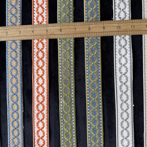 May include: Seven vertical strips of decorative woven trim featuring a repeating laurel wreath pattern. A wooden ruler is placed horizontally across the top, showing the strips measure approximately 13 inches in length. Colors include teal, tan, orange, green, blue, white, and cream.