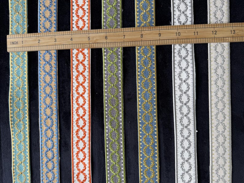 May include: Seven vertical strips of decorative woven trim featuring a repeating laurel wreath pattern. A wooden ruler is placed horizontally across the top, showing the strips measure approximately 13 inches in length. Colors include teal, tan, orange, green, blue, white, and cream.