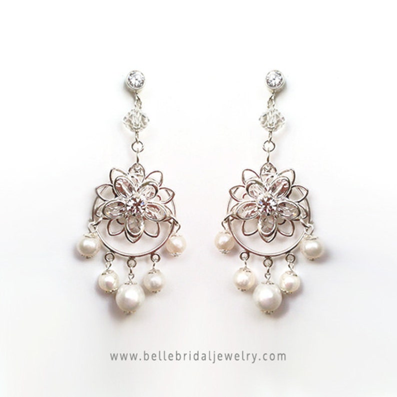 Puede incluir: Pendientes de aro de plata con un dise&ntilde;o floral y detalles de perlas. Los pendientes presentan una delicada flor calada con un centro de cristal transparente y una fila de perlas que cuelgan debajo.