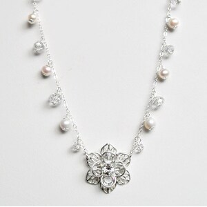 Puede incluir: Un collar de plata con un delicado colgante de flor y detalles de cristal y perlas.