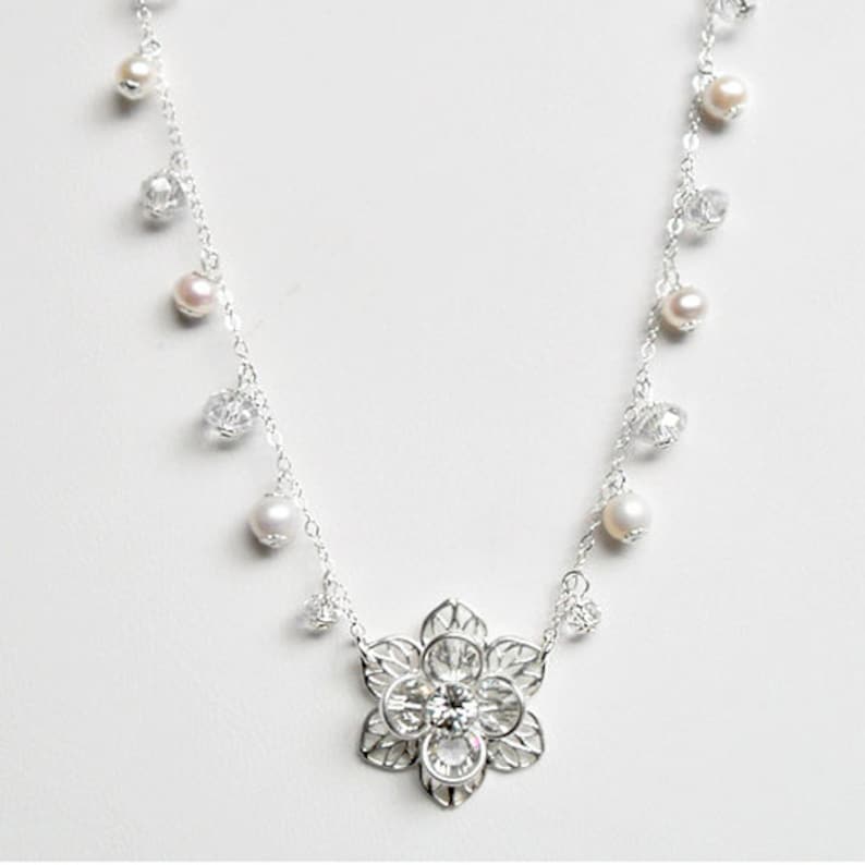 Puede incluir: Un collar de plata con un delicado colgante de flor y detalles de cristal y perlas.