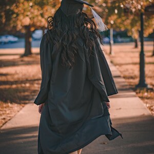 May include: A person wearing a black graduation gown and cap walks away from the camera on a paved path. The person's hair is long and dark, and the gown is flowing in the wind.