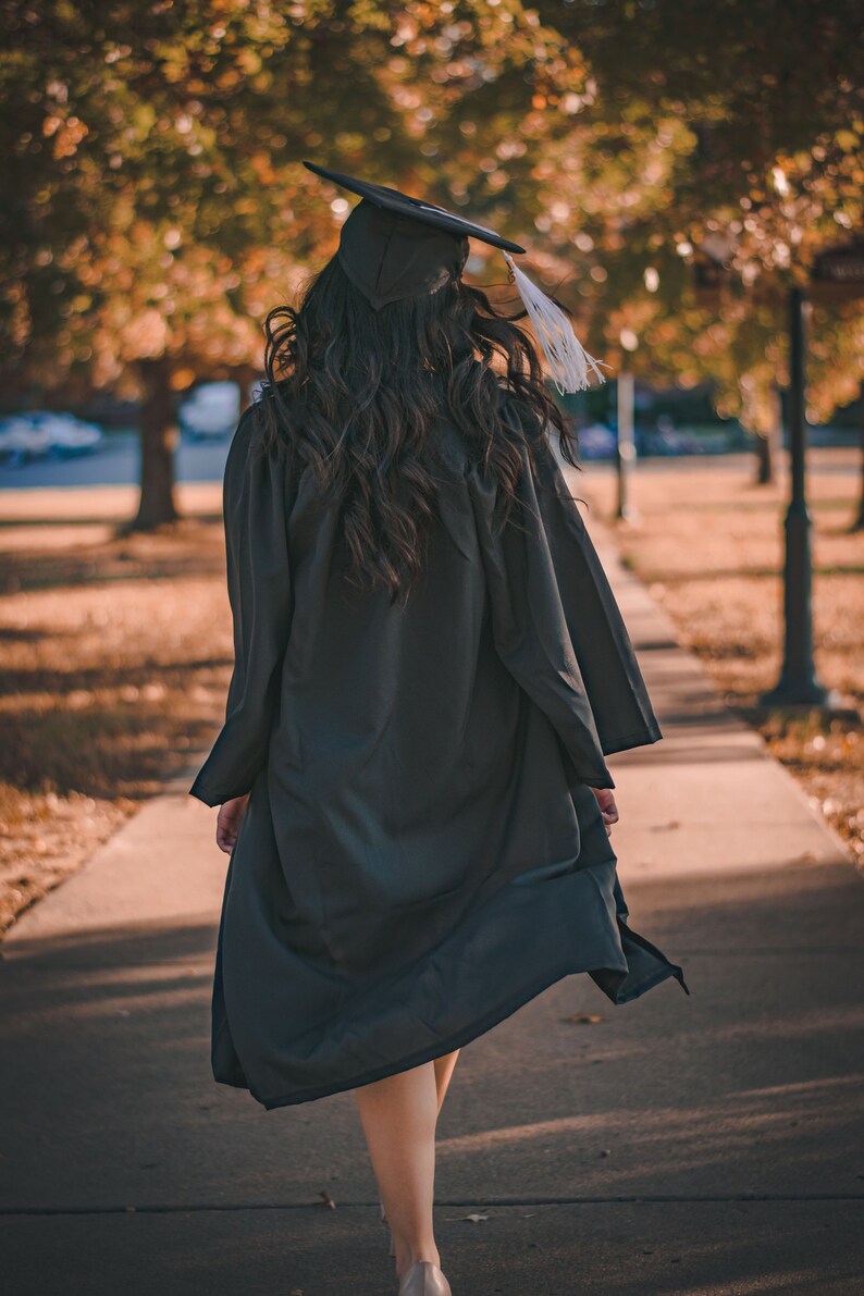 May include: A person wearing a black graduation gown and cap walks away from the camera on a paved path. The person's hair is long and dark, and the gown is flowing in the wind.