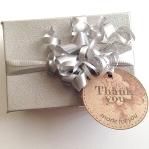 Puede incluir: Una caja de regalo plateada con una cinta y un lazo plateados. Una etiqueta marr&oacute;n con el texto "Gracias hecho para ti" est&aacute; unida a la caja.