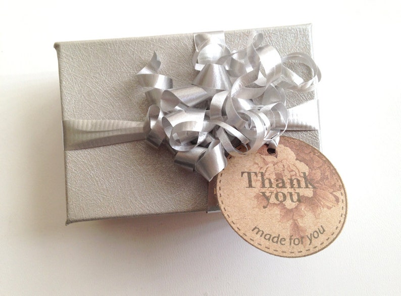 Puede incluir: Una caja de regalo plateada con una cinta y un lazo plateados. Una etiqueta marr&oacute;n con el texto "Gracias hecho para ti" est&aacute; unida a la caja.