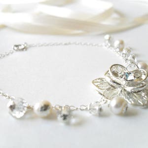 Puede incluir: Un brazalete de cadena de plata con un delicado dise&ntilde;o floral y detalles de perlas. El brazalete presenta un gran dije de flor intrincado con un centro de cristal transparente.