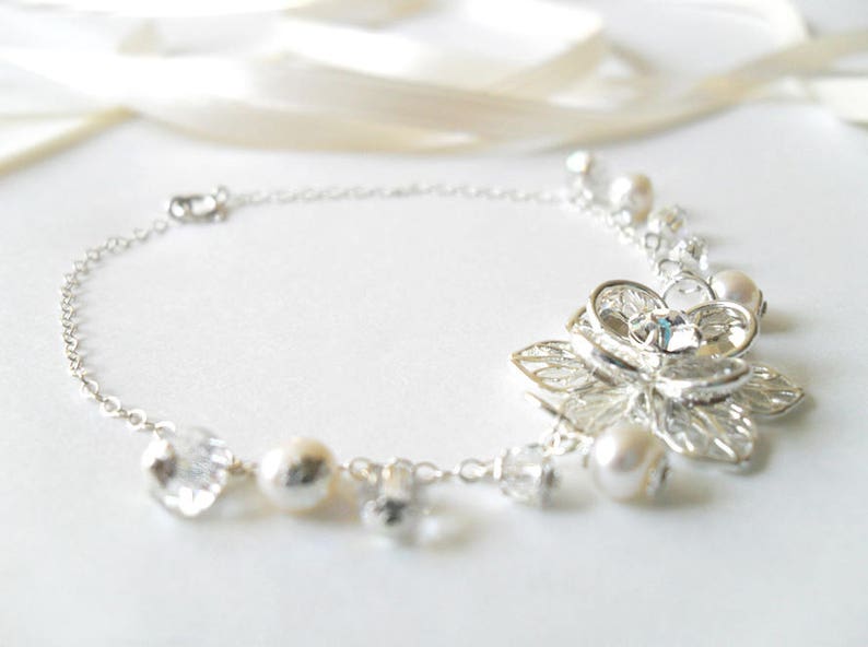 Puede incluir: Un brazalete de cadena de plata con un delicado dise&ntilde;o floral y detalles de perlas. El brazalete presenta un gran dije de flor intrincado con un centro de cristal transparente.