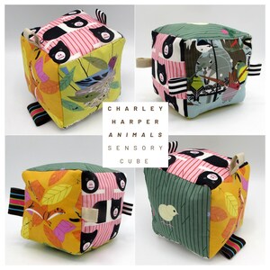 May include: A Charley Harper Animals Sensory Cube, a soft fabric cube toy with various animal and nature-themed patterns. The cube features a mix of colors, including yellow, pink, green, and black, with attached fabric tags for sensory exploration.