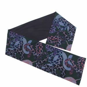 May include: A long, dark scarf with a unique pattern. The scarf features a dark green background with a repeating design of snakes in shades of blue, pink, and white. The back of the scarf is solid black.