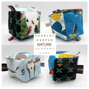 May include: A Charley Harper Nature Sensory Cube featuring various nature-themed fabric panels. The cube has multiple colors, including blue, green, and white, with illustrations of birds, leaves, and deer. Each side has a different design, and it includes fabric loops.