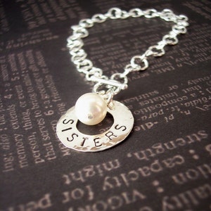 May include: A silver charm bracelet with a round charm that says "SISTERS" and a white pearl charm. The bracelet is on a dark background.