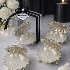 May include: Clam shell-shaped candles with pearl accents. The candles are white and sit in silver-colored holders. One candle is packaged in a clear box with a black ribbon and a label that reads "MIA & LEO 11.02.2026".